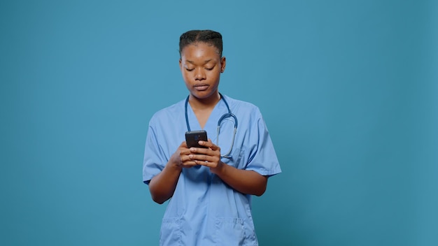 Afro-Amerikaanse vrouw in uniform die smartphone gebruikt om gezondheidszorg en medicijnen in de studio te oefenen. Medisch assistent met stethoscoop die naar het scherm van de mobiele telefoon kijkt om ziekteonderzoek te doen