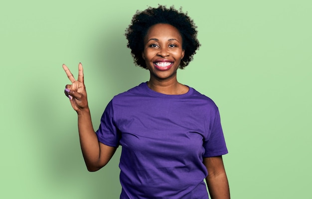 Afro-Amerikaanse vrouw met afrohaar in casual paars t-shirt glimlachend kijkend naar de camera met vingers die overwinningsteken nummer twee doen