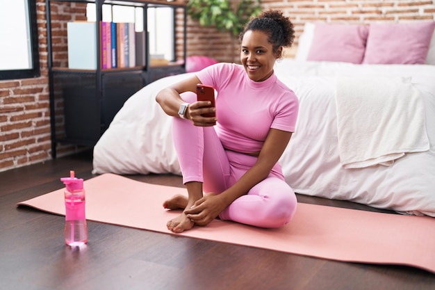 Afro-Amerikaanse vrouw met behulp van smartphone zittend op yoga mat in de slaapkamer