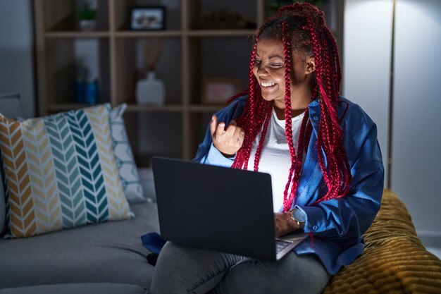 Afro-amerikaanse vrouw met gevlochten haar die 's nachts een computerlaptop gebruikt, wenkend kom hier gebaar met de hand uitnodigend blij en glimlachend verwelkomend
