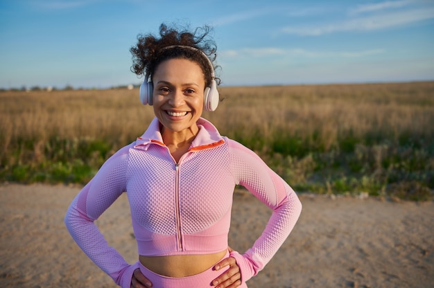 Afro-Amerikaanse vrouw met koptelefoon die haar handen op een taille legt, glimlachend met een brede glimlach