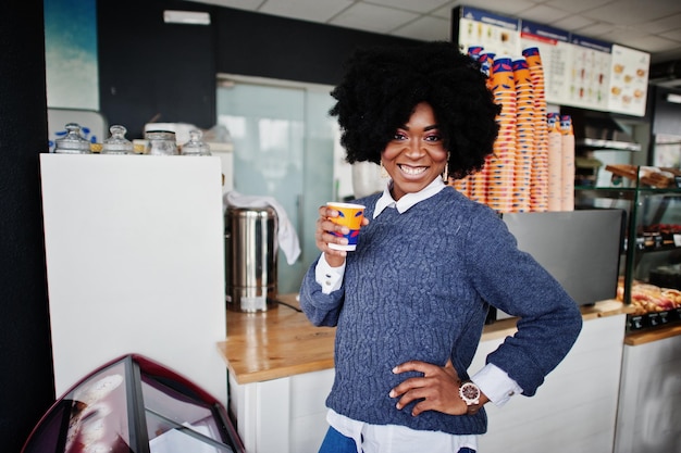 Afro-Amerikaanse vrouw met krullend haar draagt een trui in café binnen met een kopje thee of koffie