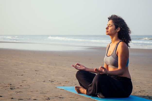 Afro-amerikaanse vrouw met krullend haar en gesloten ogen die diep ademhalen en zichzelf kalmeren op een leeg ochtendstrand na surya namaskar