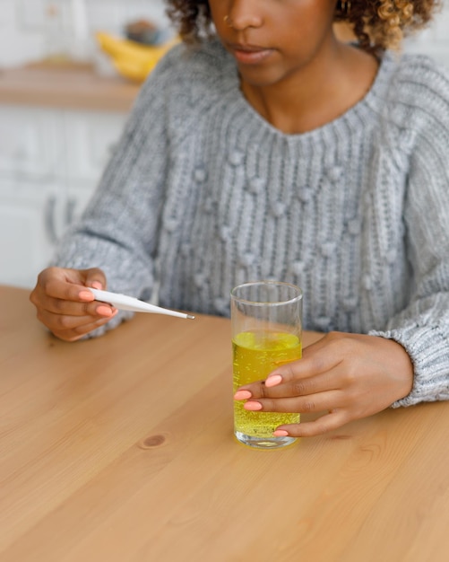 Afro-Amerikaanse vrouw met verkoudheid meet haar lichaamstemperatuur en drinkt oplosbare pillen of vitamines Een vermoeide zieke vrouw