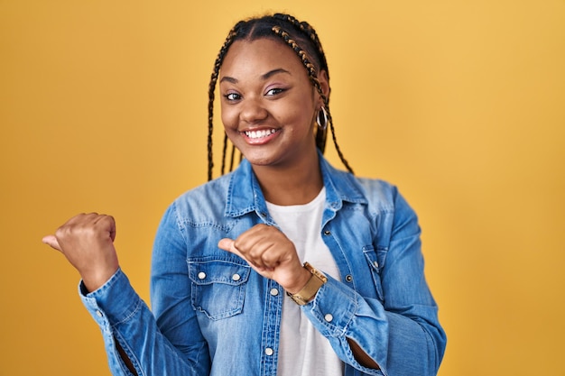 Afro-Amerikaanse vrouw met vlechten die over een gele achtergrond staan, wijzend naar achteren met hand en duimen omhoog glimlachend zelfverzekerd