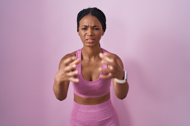 Afro-amerikaanse vrouw met vlechten die sportkleding dragen over roze achtergrond schreeuwen gefrustreerd van woede, handen proberen te wurgen, gek schreeuwen
