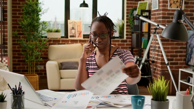 Afro-Amerikaanse vrouw praten over mobiel telefoongesprek thuis, op afstand werken op laptop. Mensen op smartphone bellen om te discussiëren over telefoonlijn, telewerkbaan en telecommunicatie.