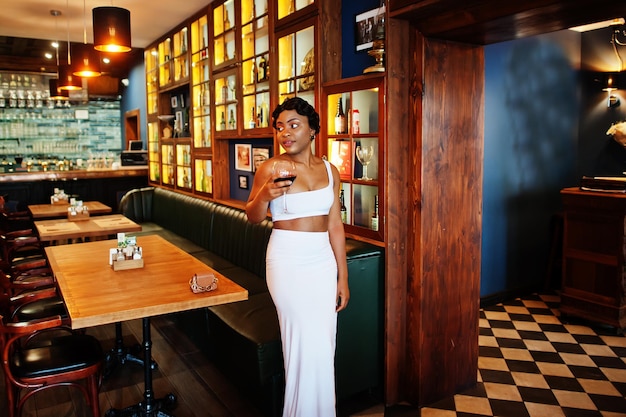 Afro-Amerikaanse vrouw, retro kapsel in witte jurk in restaurant met glas wijn.