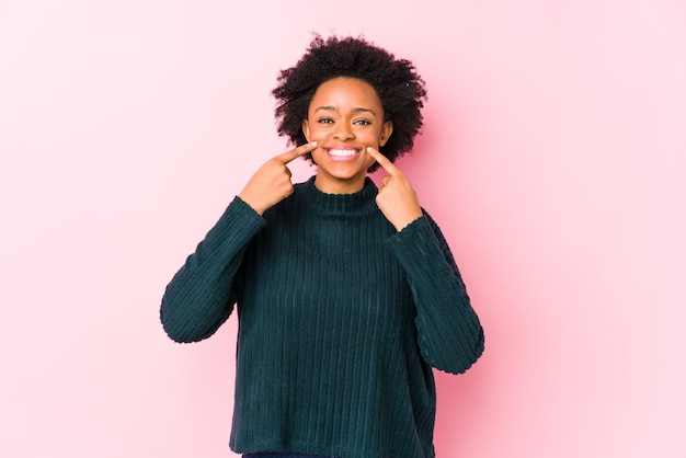 Afro-Amerikaanse vrouw van middelbare leeftijd tegen een roze muur geïsoleerde glimlach, wijzende vingers naar de mond.