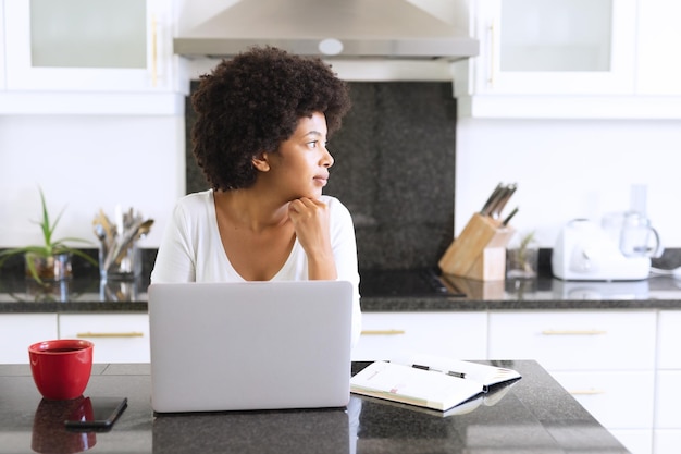 Afro-Amerikaanse vrouw zit in de keuken met behulp van laptop en denkt