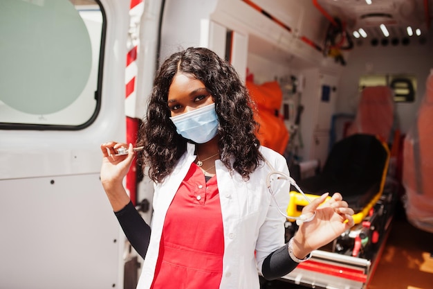 Afro-amerikaanse vrouwelijke paramedicus in gezichtsbeschermend medisch masker dat voor de ambulanceauto staat.