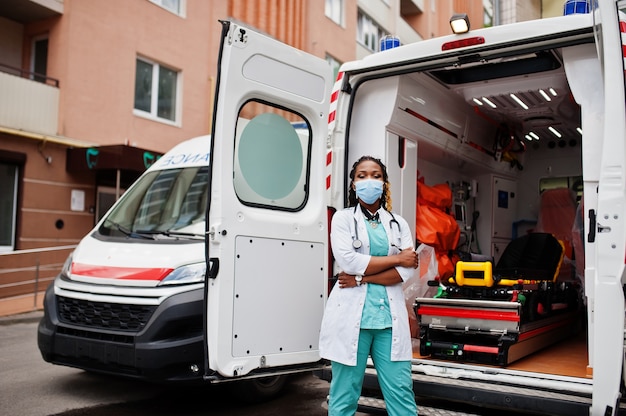 Foto afro-amerikaanse vrouwelijke paramedicus in gezichtsmasker staande voor ambulance auto