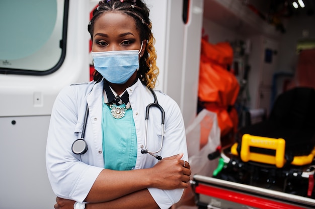 Afro-Amerikaanse vrouwelijke paramedicus in gezichtsmasker staande voor ambulance auto