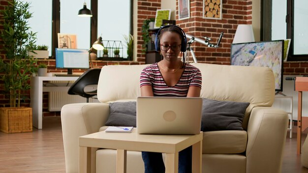 Afro-amerikaanse werknemer werkt op afstand op laptop, luistert naar muziek op een koptelefoon en zit op de bank. zakenvrouw surfen op internet voor externe baan thuis, online netwerk.