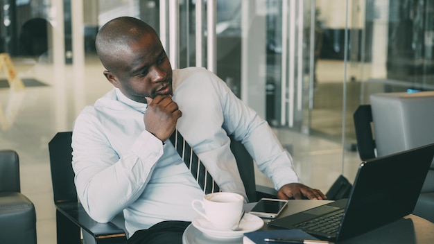 Afro-Amerikaanse zakenman in formele kleding die naar zijn laptop kijkt en nadenkt over zijn businessplan in een stijlvol café tijdens de lunchpauze Hij is attent en denkt aan een project