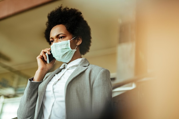 Afro-Amerikaanse zakenvrouw die op een smartphone praat terwijl ze door de luchthavengang loopt met een beschermend masker op haar gezicht Ruimte kopiëren