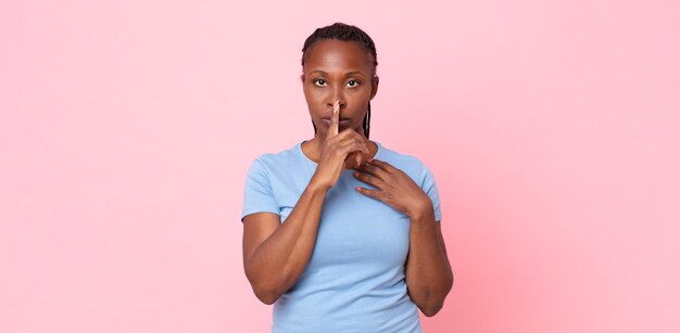 Afro black adult woman looking serious and cross with finger pressed to lips demanding silence or quiet, keeping a secret