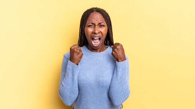 Afro black pretty woman shouting aggressively with annoyed, frustrated, angry look and tight fists, feeling furious