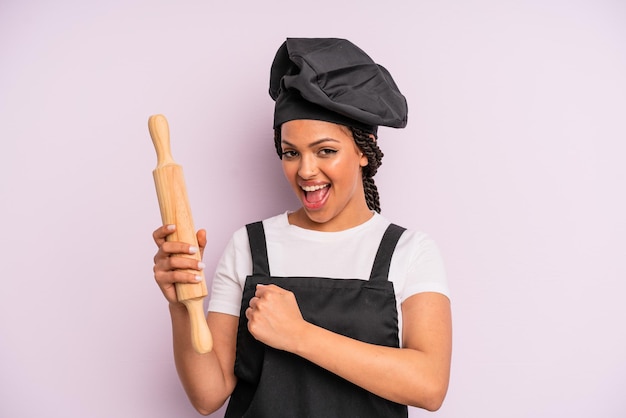 Afro black woman feeling happy and facing a challenge or\
celebrating. chef with a roller pin