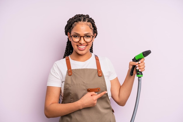 Afro black woman smiling cheerfully, feeling happy and pointing to the side. gardener hose concept