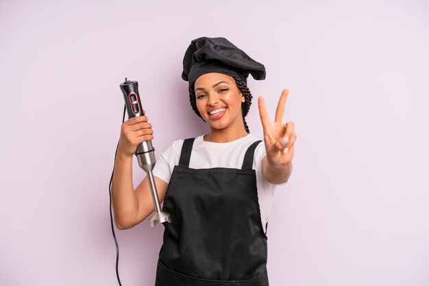 afro black woman smiling and looking friendly, showing number two. chef with hand blender