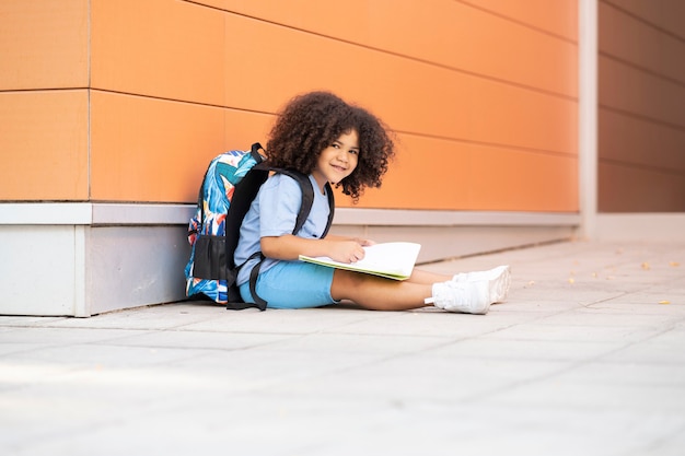 Afro boy with his backpack on is sitting writing in a notebook