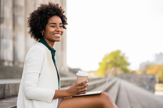 Afro business woman using her laptop while sitting on stairs outdoors. Urban and business concept.