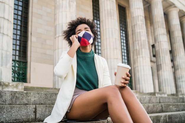 Afro business woman wearing protective mask and talking on the phone while sitting on stairs outdoors at the street. Business and urban concept.