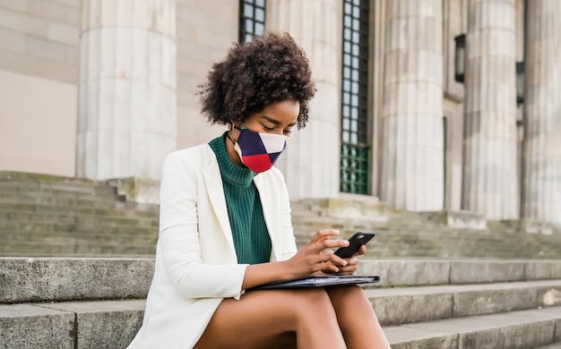 Donna d'affari afro indossando maschera protettiva e utilizzando il suo telefono cellulare