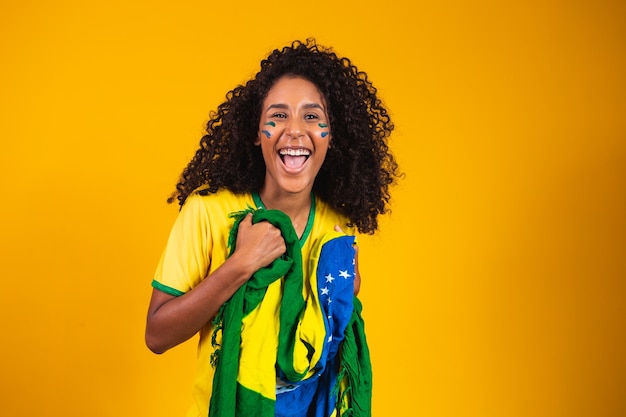 Afro girl cheering for favorite brazilian team, holding national flag in yellow background.