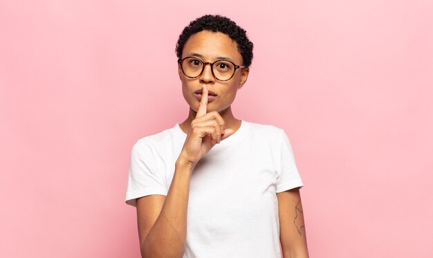 Afro jonge zwarte vrouw die ernstig en kruis kijkt met de vinger tegen de lippen gedrukt en stilte of stilte eist, een geheim bewaren