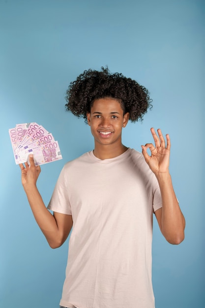 Photo afro-latino young man holding euro banknotes making good sign with fingers, friendly gesture smiling excellent symbol