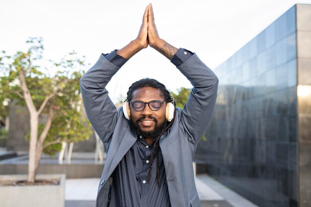 Afro man in pak en met bril doet yoga buiten terwijl hij naar muziek luistert met een koptelefoon