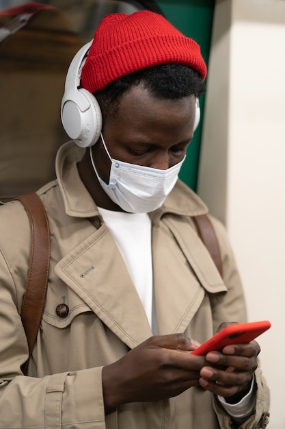 Afro man in subway, wear face mask, using mobile, listens to music