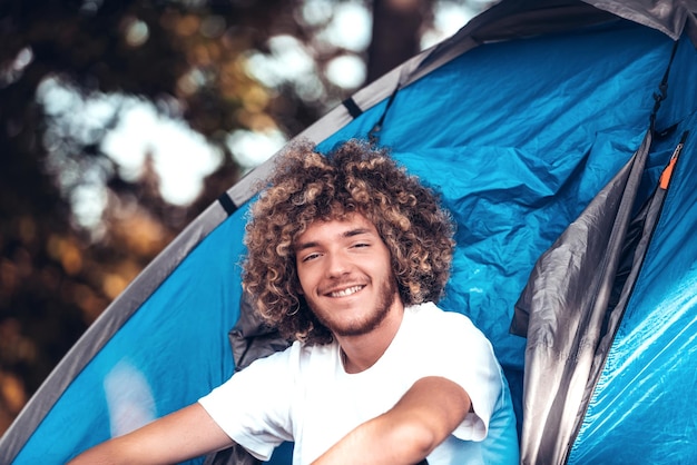 An afro teenager is sitting in front of a mountain tent in the early morning