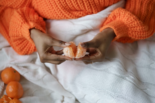 Afro vrouw handen peeling rijpe zoete mandarijn, dragen oranje trui, liggend in bed onder de plaid