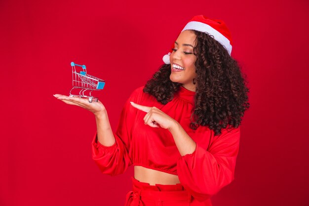 Afro vrouw in kerstmuts kerstinkopen doen. Ze laat een minikarretje zien