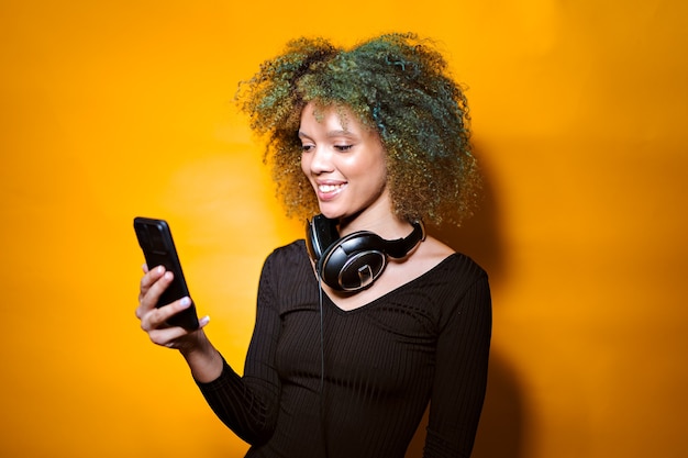 Afro-vrouw met mobiele telefoon en koptelefoon op gele achtergrond