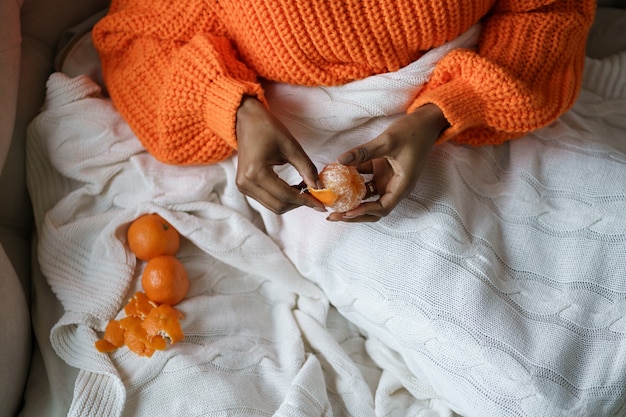 Afro woman hands peeling ripe sweet tangerine, wear orange sweater, lying in bed under the plaid