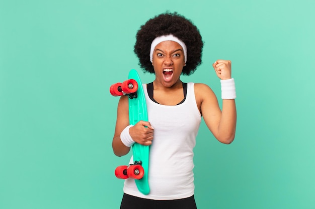 Afro woman shouting aggressively with an angry expression or with fists clenched celebrating success. skateboard concept