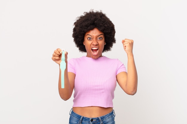 Afro woman shouting aggressively with an angry expression or with fists clenched celebrating success watch concept