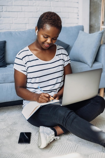 Афро-женщина учится удаленно, посещает онлайн-конференцию и делает записи в блокноте, сидя...