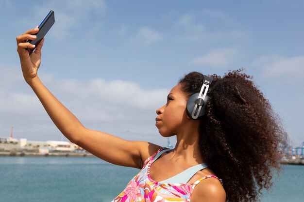 背景の海の景色の中で携帯電話でselfieを取るヘッドフォンを持つアフロ女性