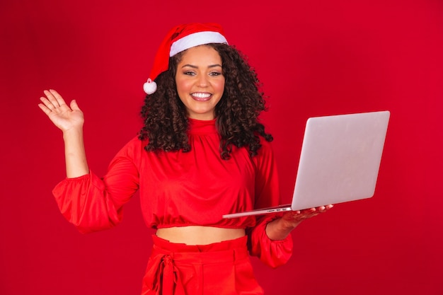 빨간색 배경에 노트북을 사용하여 산타 클로스 모자와 아프리카 여자.