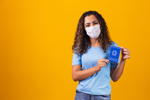 Afro woman with surgical mask holding brazilian work card on yellow background. Work, economy and pandemic concept