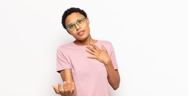 Afro young black woman feeling happy and in love, smiling with one hand next to heart and the other stretched up front