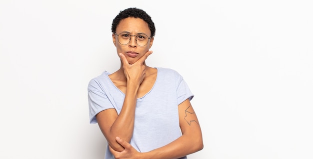 Afro young black woman looking serious, thoughtful and distrustful, with one arm crossed and hand on chin, weighting options