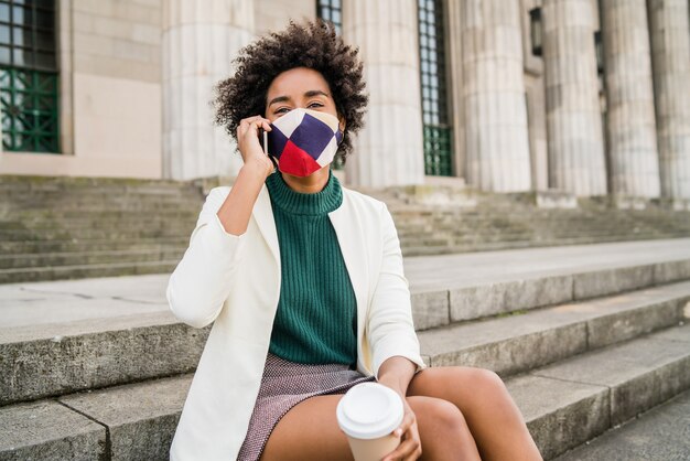 Afro zakenvrouw beschermend masker dragen en praten aan de telefoon zittend op de trap buiten op straat. Bedrijfs- en stedelijk concept.