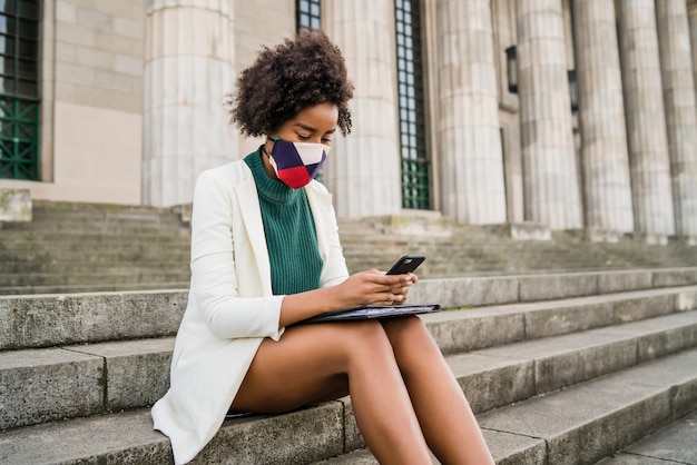 Afro zakenvrouw dragen beschermend masker en met behulp van haar mobiele telefoon zittend op de trap buiten op straat. Bedrijfs- en stedelijk concept.