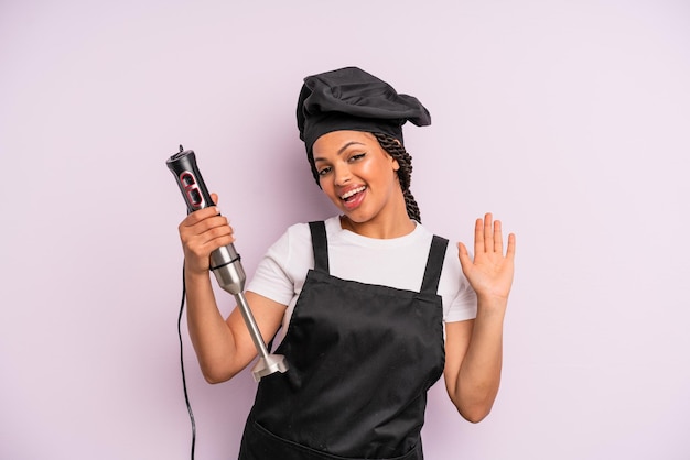 Afro-zwarte vrouw die vrolijk lacht, met de hand zwaait, je verwelkomt en begroet. chef-kok met staafmixer